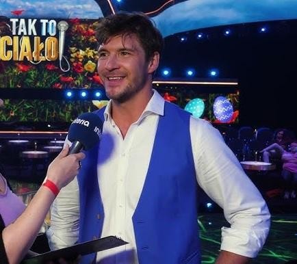 Mikołaj Roznerski na planie filmowym, w swobodnej stylizacji, w tle widoczne elementy sprzętu filmowego.