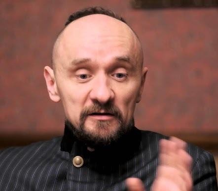 Jan Klata w dynamicznej scenie z próby teatralnej, z intensywnym wyrazem skupienia, otoczony elementami scenografii i oświetleniem teatralnym.
