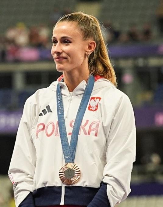 Natalia Bukowiecka na bieżni, celebrująca zwycięstwo z medalem na szyi, uśmiechnięta i triumfująca.