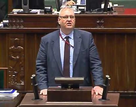 Bogdan Roman Święczkowski podczas wystąpienia publicznego, z dynamiczną mimiką, w tle rozmyty budynek rządowy.