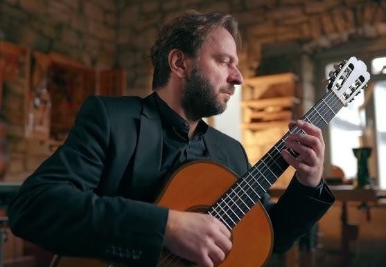 Marcin Dylla w zamyśleniu, z subtelnie widoczną gitarą klasyczną w tle, symbolizującą jego edukację i poświęcenie muzyce.