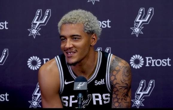Jeremy Sochan z unikalną fryzurą i skupionym wyrazem twarzy, w koszulce San Antonio Spurs, na tle boiska do koszykówki.