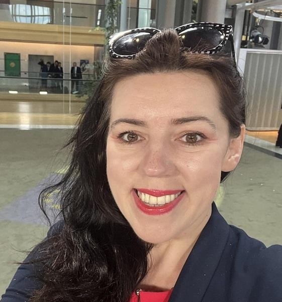 Dominika Ćosić w dynamicznej scenie pracy dziennikarskiej, np. podczas konferencji prasowej lub pisania artykułu, z tłem nawiązującym do europejskiego środowiska mediów.