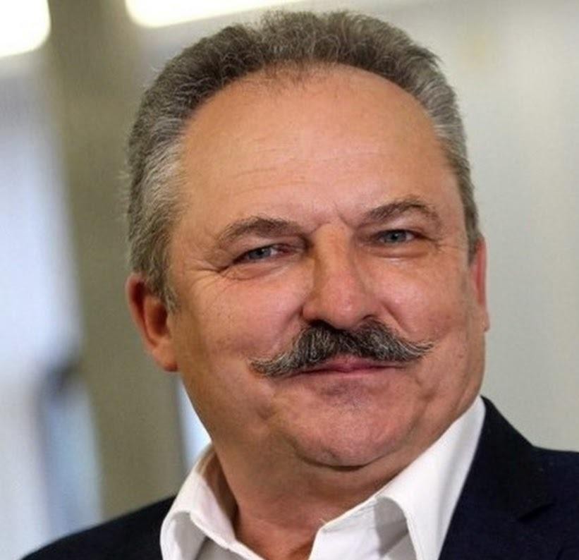 Marek Jakubiak w dynamicznej pozie, być może podczas wystąpienia publicznego, ubrany w elegancki strój, z lekko rozmytym tłem sugerującym wydarzenie.