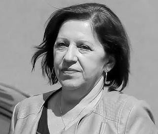 Barbara Skrzypek podczas przesłuchania w prokuraturze, siedząca przy stole z dokumentami, w tle widoczni mężczyźni, co symbolizuje kluczowe wydarzenie związane ze śledztwem w sprawie spółki Srebrna.