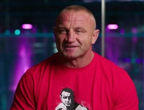 Mariusz Pudzianowski, z widocznymi mięśniami, w dynamicznej pozie, na tle symbolizującym sporty siłowe i MMA.