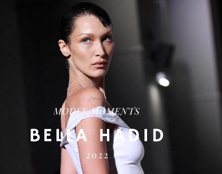Bella Hadid za kulisami pokazu mody, uchwycona w naturalnym momencie, otoczona elementami świadczącymi o intensywności pracy w świecie mody.
