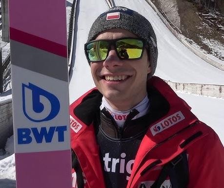 Jakub Wolny w kombinezonie skoczka narciarskiego, zdeterminowany wyraz twarzy, w tle rozmazana skocznia.